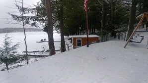 A cabin with a snow-covered deck, a swing set, and a small shed in a forested area. - Family friendly lakefront cottage (Pickerel)