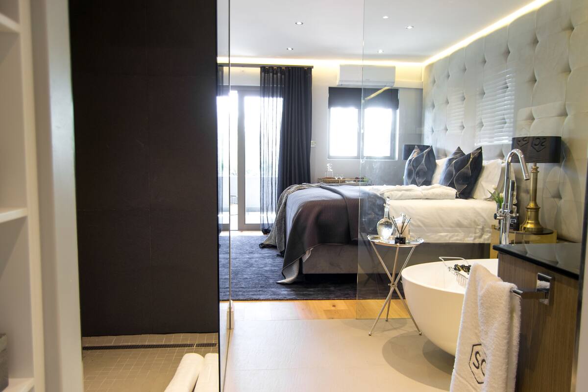 A modern hotel room with a large bed, a separate sofa, a round bathtub, and a vanity with a mirror.