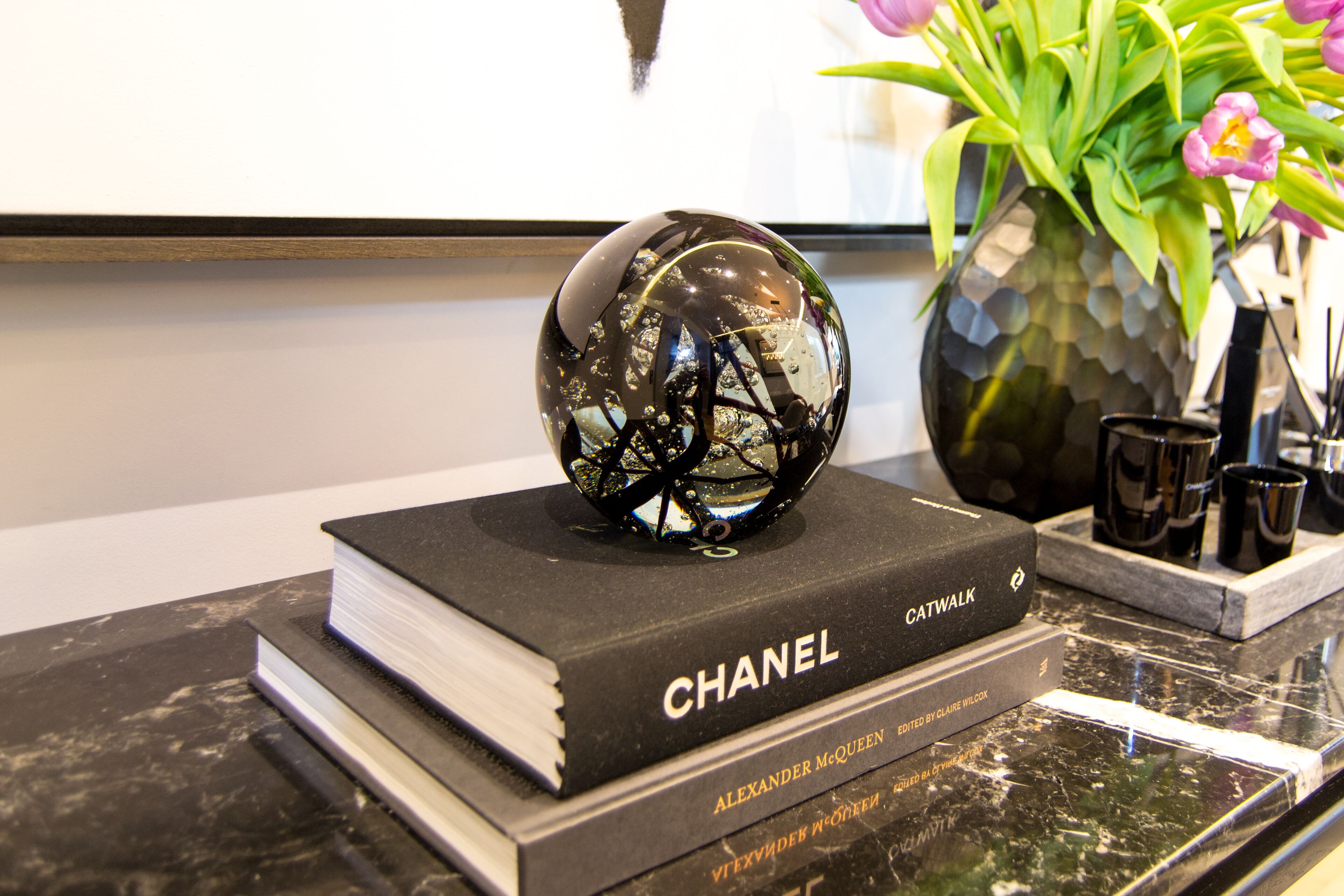 A book titled "CHANEL" on a marble surface, a decorative glass sphere, and a vase with flowers.