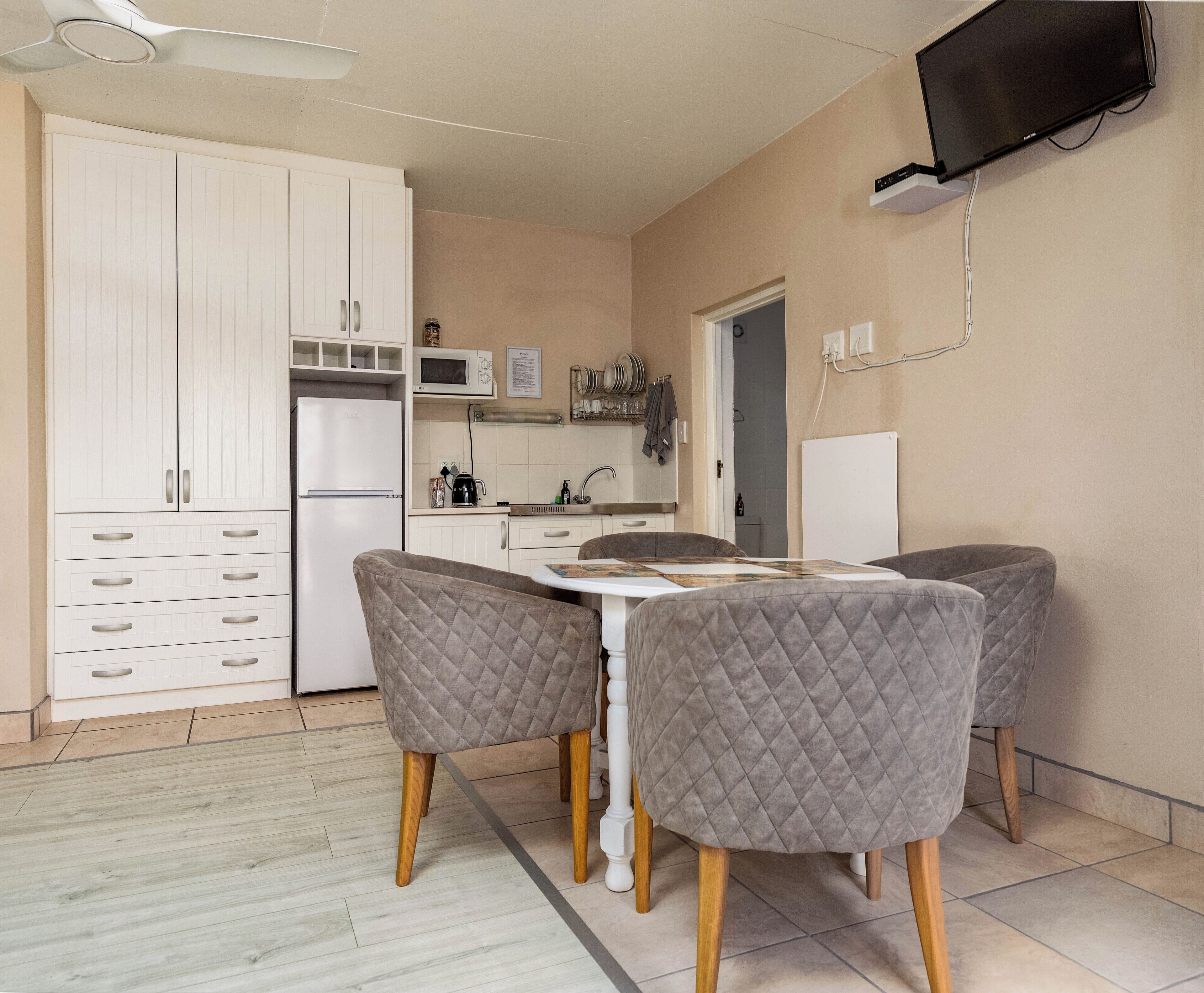 A compact kitchen with white cabinets, a microwave, and a sink. There is a dining table with four chairs and a flat-screen TV mounted on the wall.