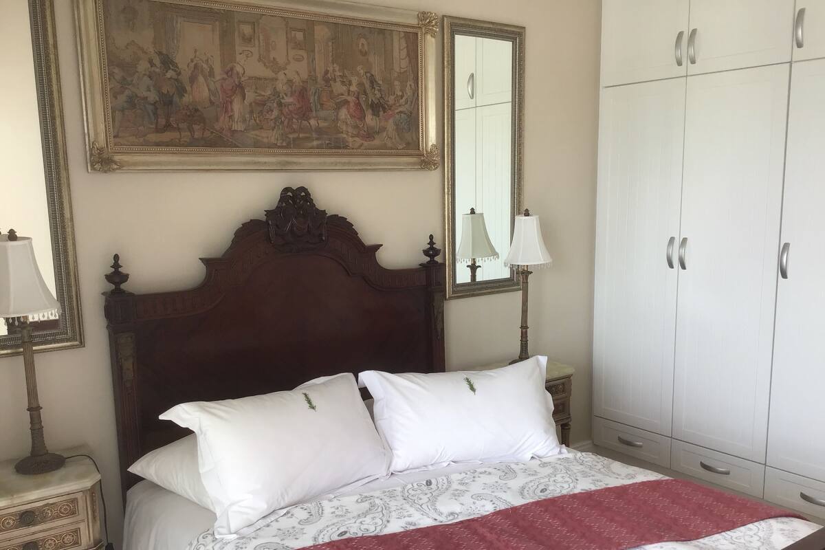 A bedroom with a wooden headboard, a bed with white linens, a red throw blanket, a nightstand with a lamp, a large mirror, and a wardrobe.