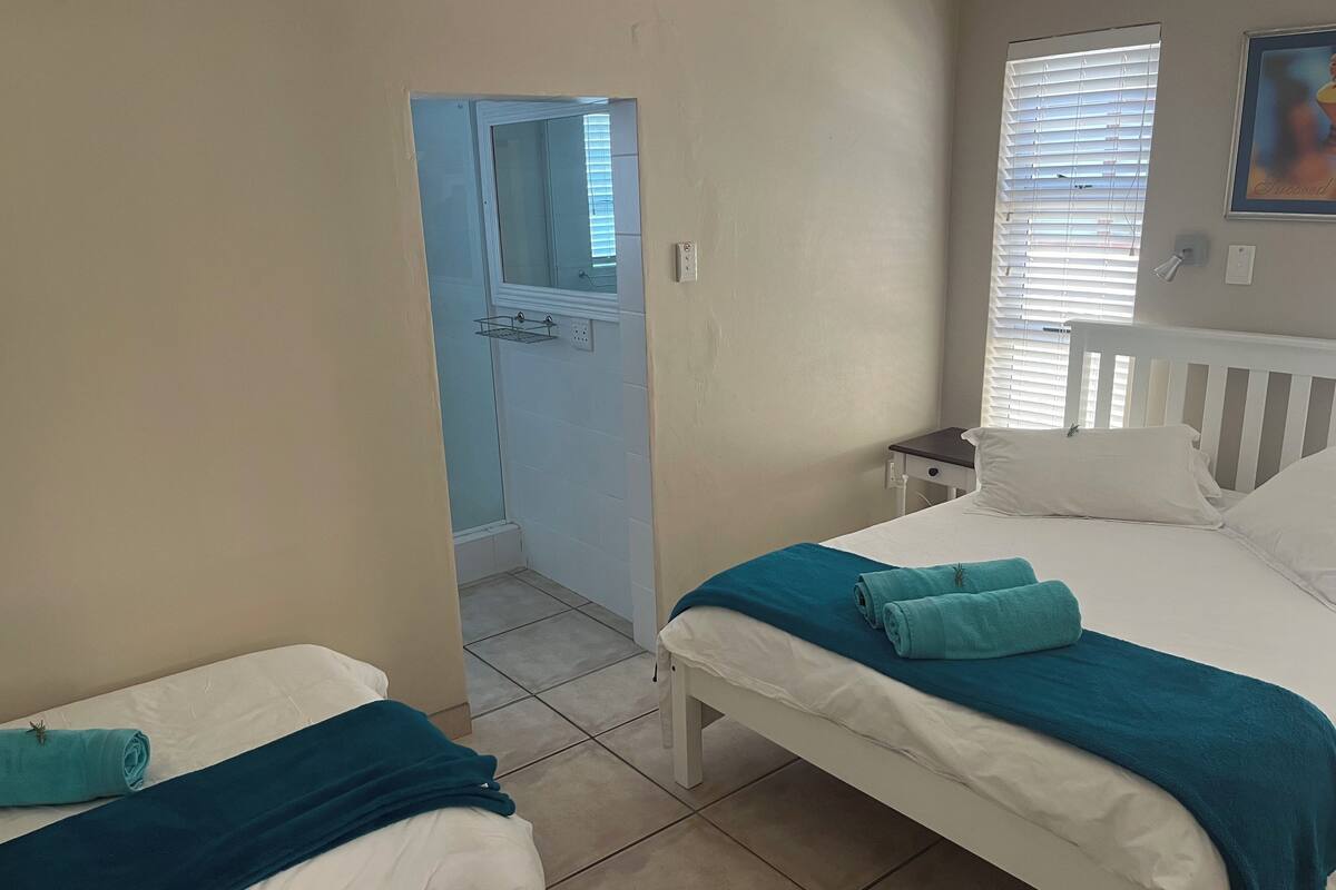 A bedroom with two beds, a small bedside table, a window with blinds, and a bathroom visible through an open door.