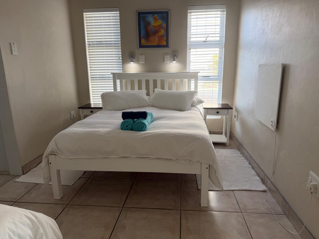 Une chambre à coucher comprenant un lit, deux tables de chevet, une fenêtre avec des stores et un luminaire fixé au mur.