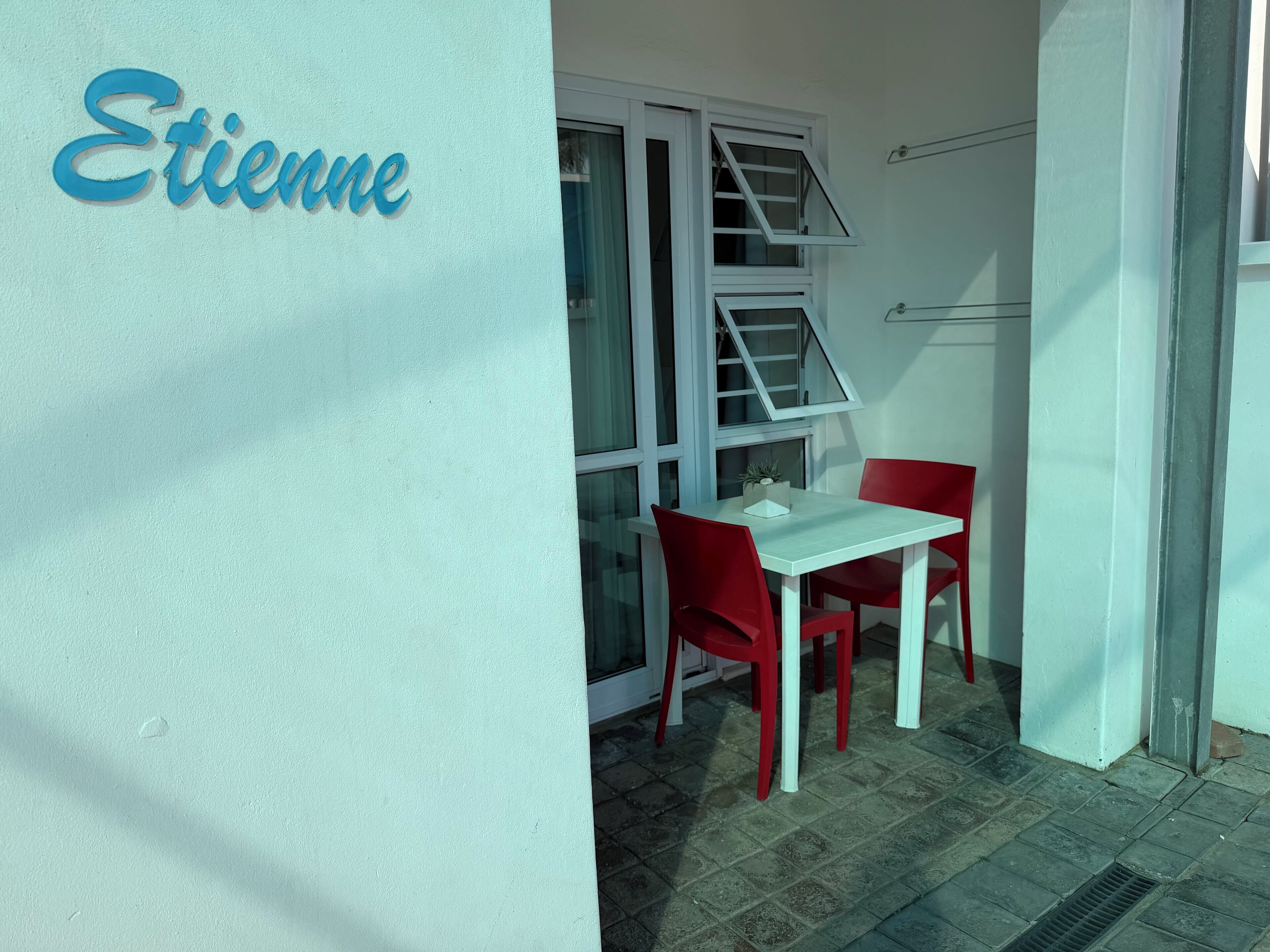 A small outdoor seating area with a white table and red chairs, a window with blinds, and a sign reading "Etienne".