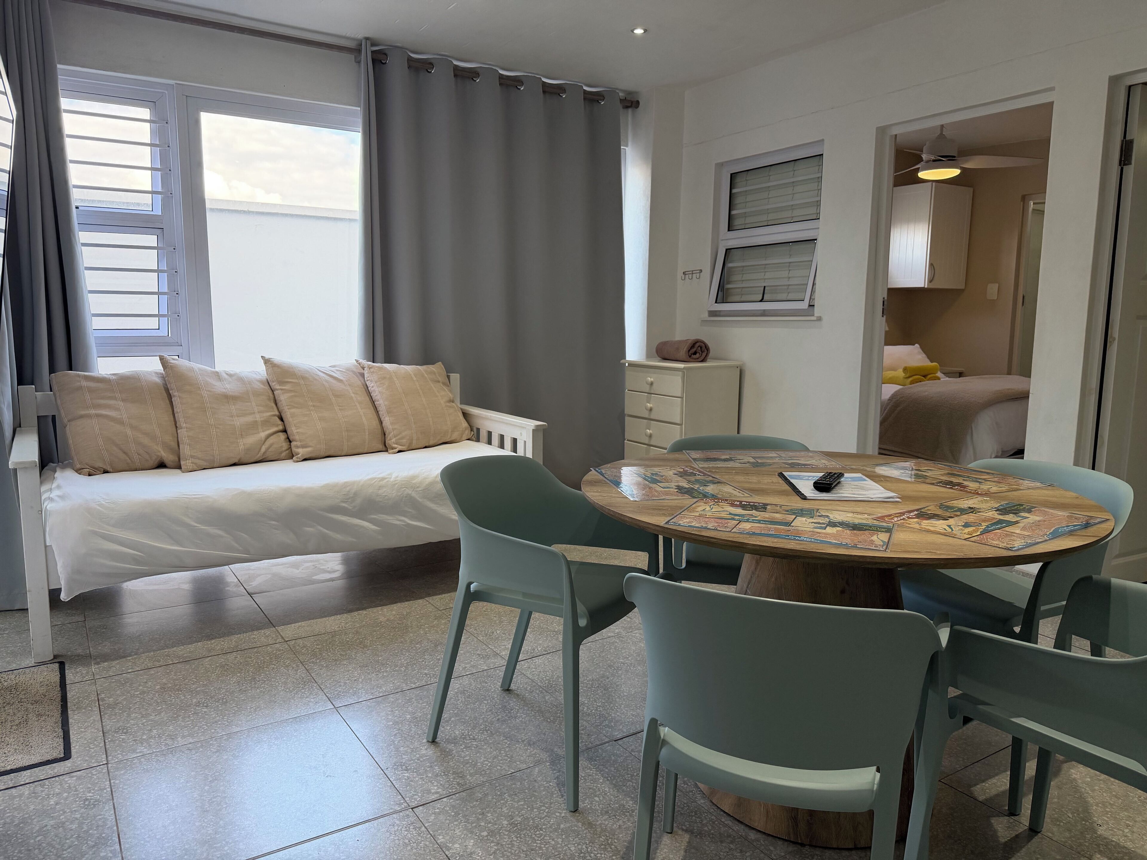 A room with a bed, a dining table with chairs, a small cabinet, and a window with curtains.