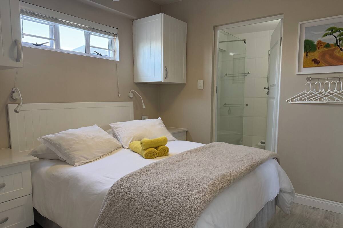 A bedroom with a bed, white curtains, a window, a white wardrobe, a glass shower door, and a painting on the wall.