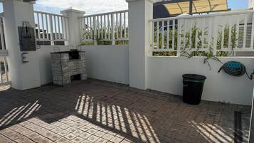 Un patio exterior con una parrilla de piedra, un bote de basura y una manguera.