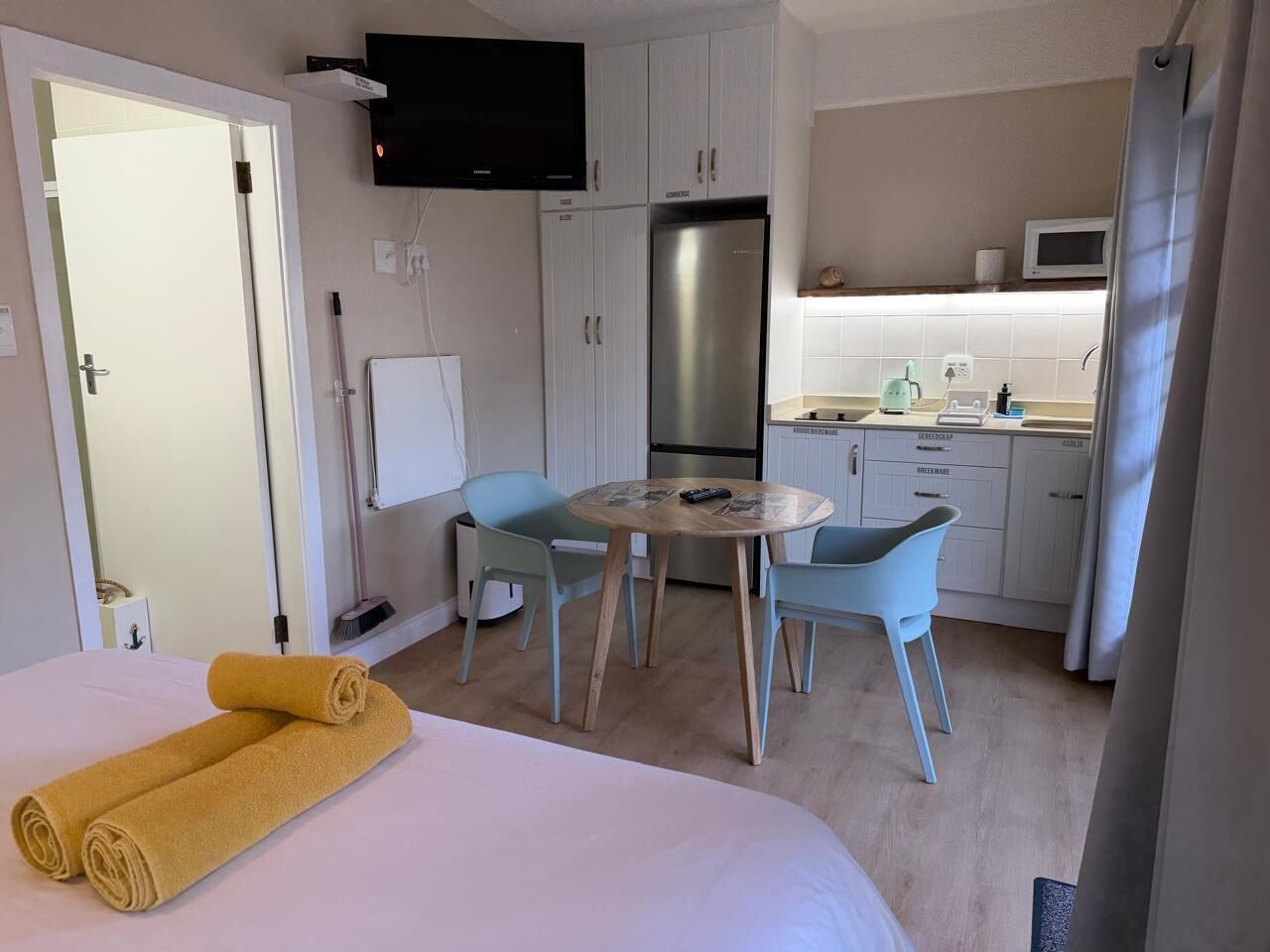 A hotel room with a bed, a round table with chairs, a kitchenette, and a TV mounted on the wall.