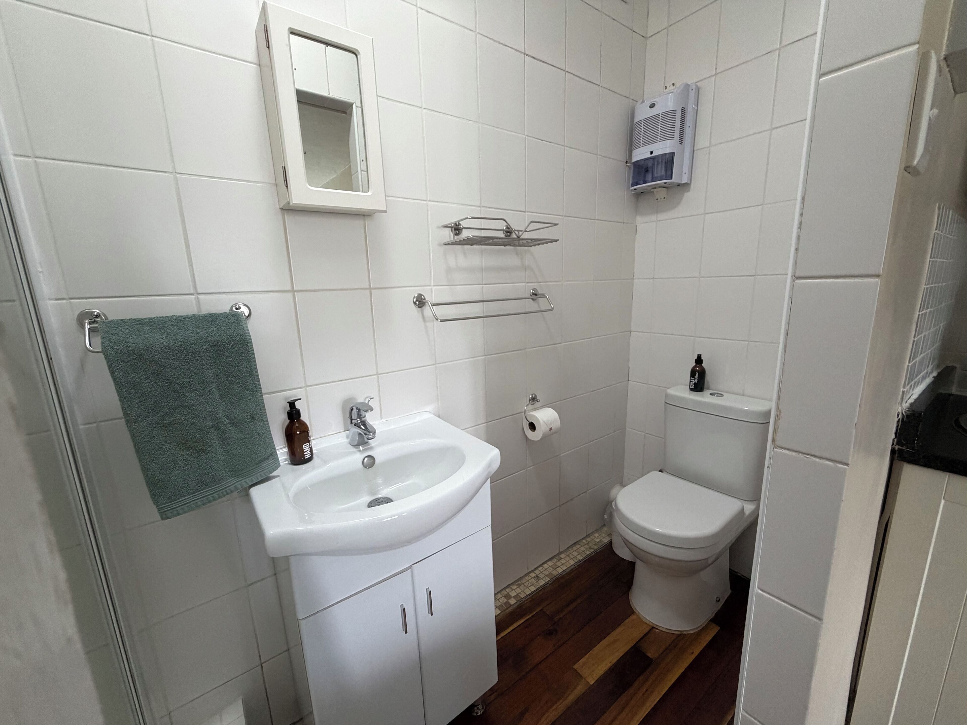 A bathroom with a white sink, toilet, and mirror. There is a towel rack with a green towel, a soap dispenser, and a toilet paper holder.