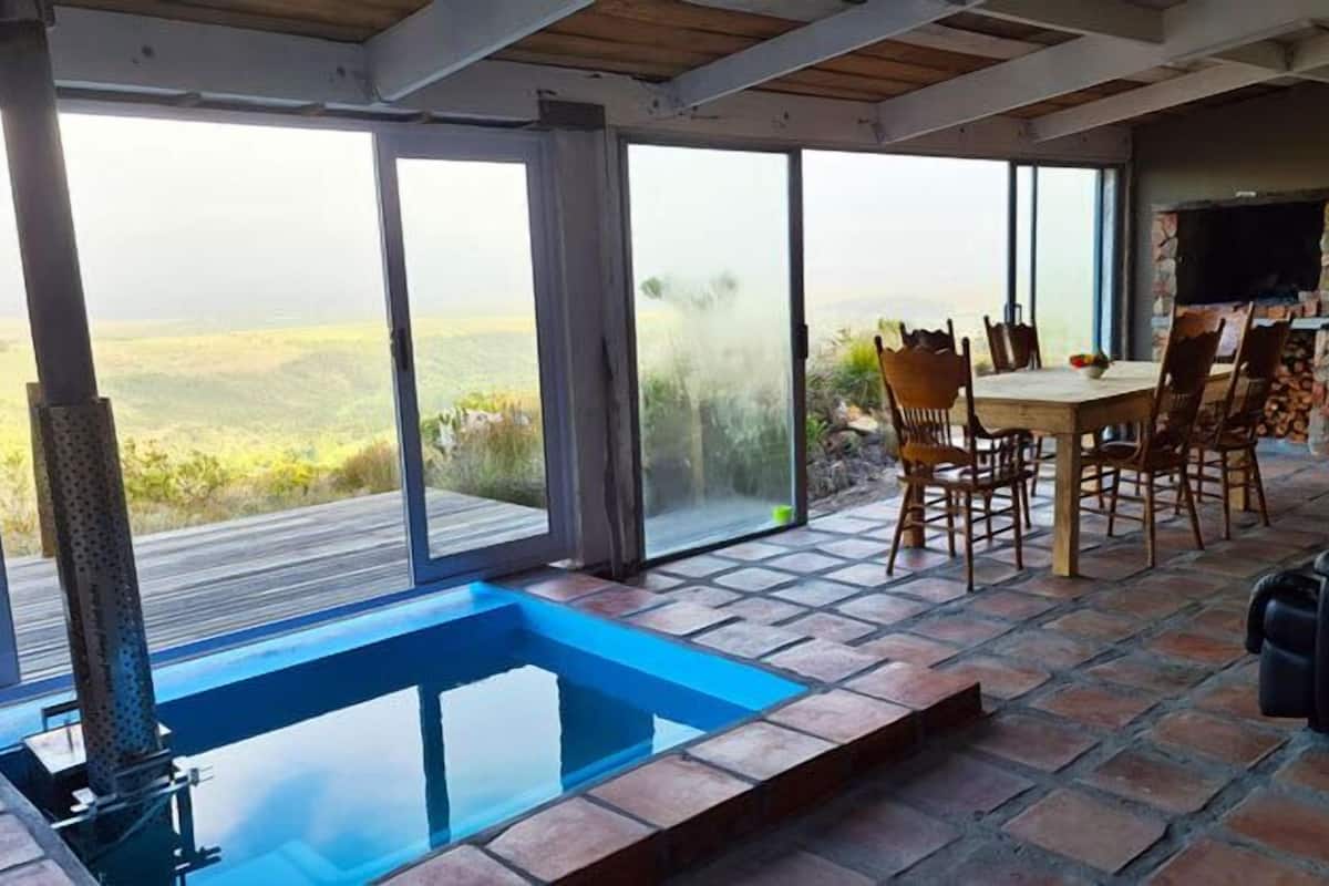 A dining area with a view of the countryside, a pool, and a stone fireplace.