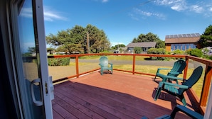 A wooden deck with two green Adirondack chairs, a railing, and a view of a residential street and houses. - Sea Break - Ocean view with Hot tub (Yachats)