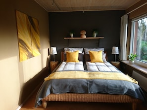 A bedroom with a large bed, two bedside tables, a window with curtains, and a wall-mounted shelf with decorative items.