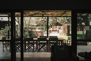 Een terras met houten tafels en stoelen, een stenen openhaard en uitzicht op een tuin en bomen.