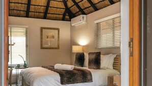 A bedroom with a thatched roof, a bed with a white comforter and a brown throw, a bedside lamp, and a framed picture on the wall. - Buccara Waterberg - Molapo (Lephalale)