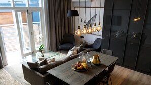 Et moderne spiseområde med et træbord, stole og en skål med frugt. En sofa og en lænestol ses i baggrunden, og en lampe hænger over bordet.