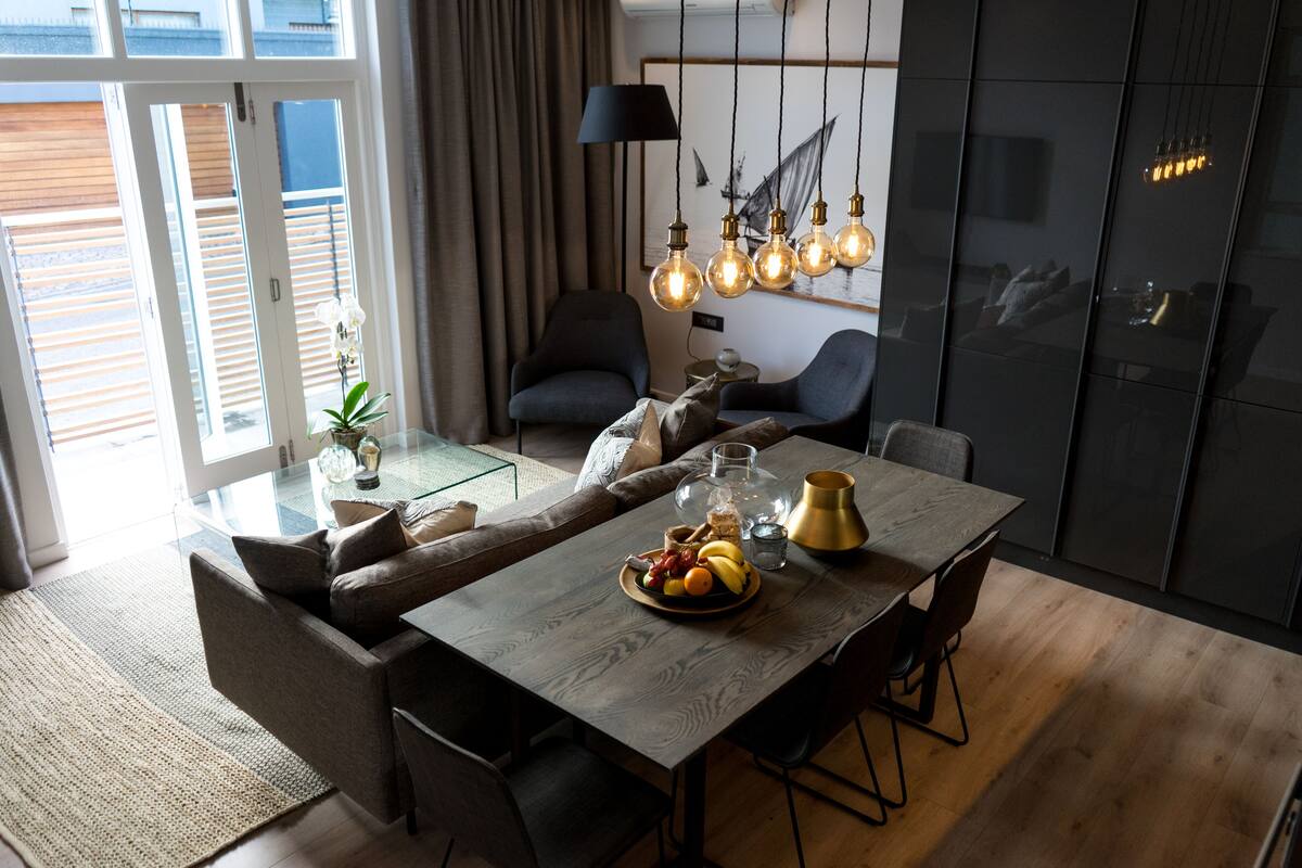 A modern dining area with a wooden table, chairs, and a fruit bowl. There is a sofa and a chair in the background, and a lamp hanging above the table.