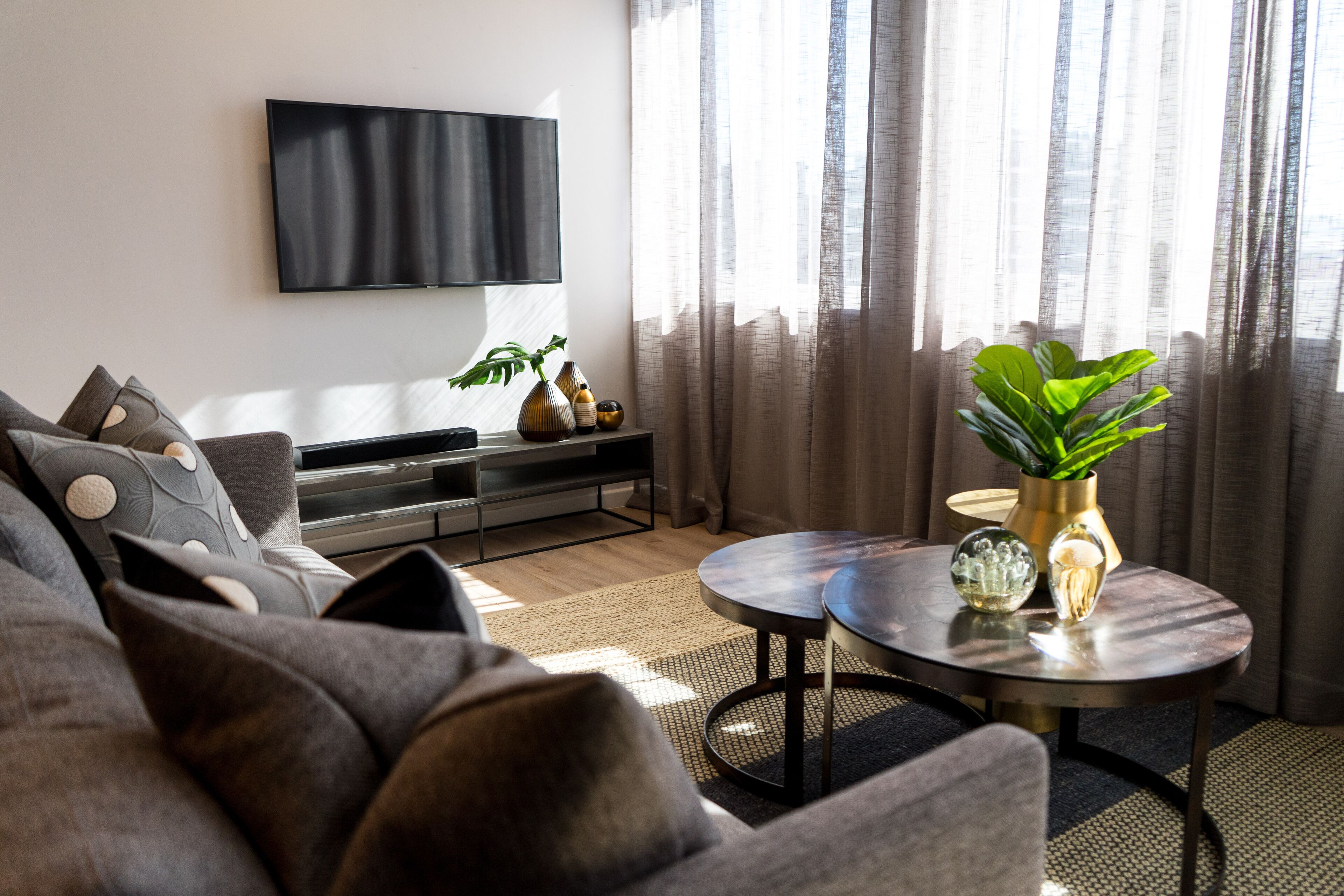 A modern living room with a flat-screen TV mounted on the wall, a round coffee table with decorative items, and a sofa with patterned cushions.