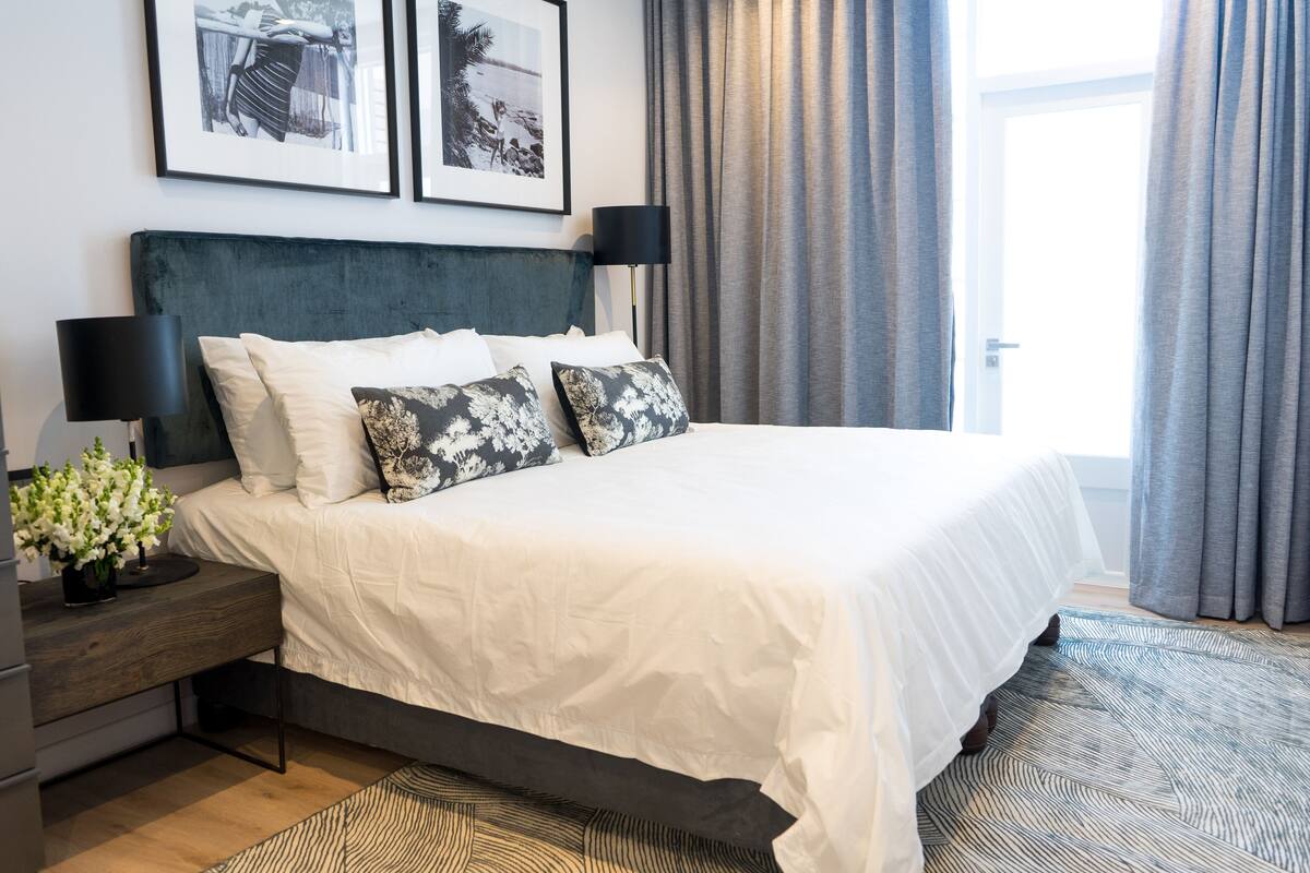 A neatly arranged bedroom with a large bed, a bedside table with a lamp, and a small plant. There are two framed pictures on the wall above the bed.