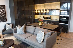 A modern living room with a grey sofa, a dining table, and a kitchen area with a large window and pendant lights. - Kayleeway Apartments (Cape Town)