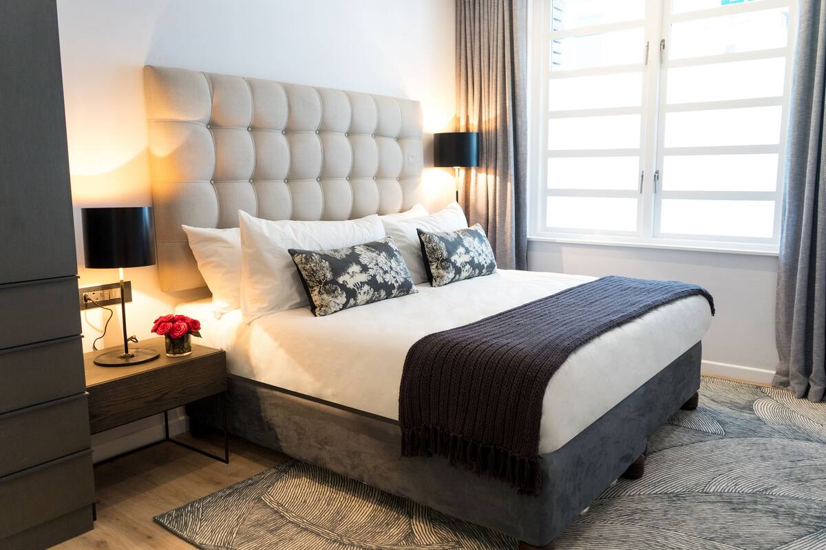 A neatly arranged bedroom with a tufted headboard, a bed with white linens and patterned pillows, a dark brown throw blanket, a bedside table with a lamp, and a decorative vase with red flowers.