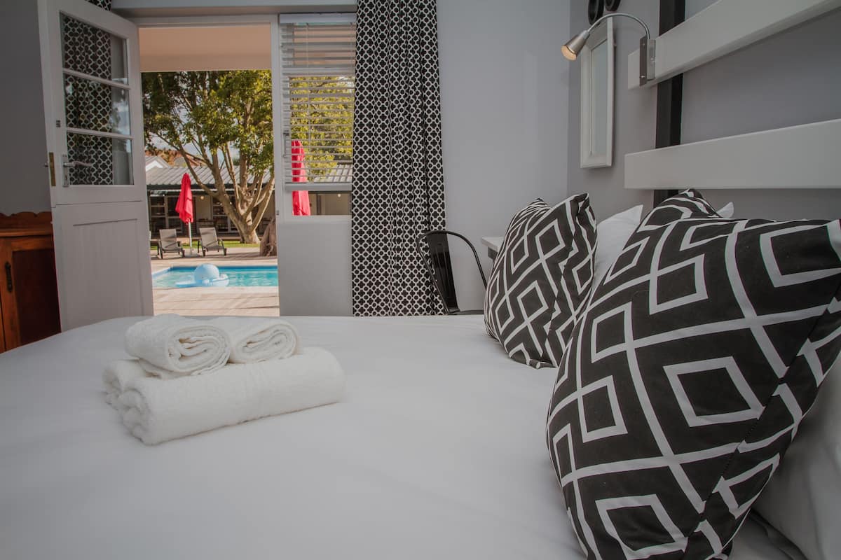 A hotel room with a bed, a window with a view of a pool and outdoor seating, and a patterned curtain.