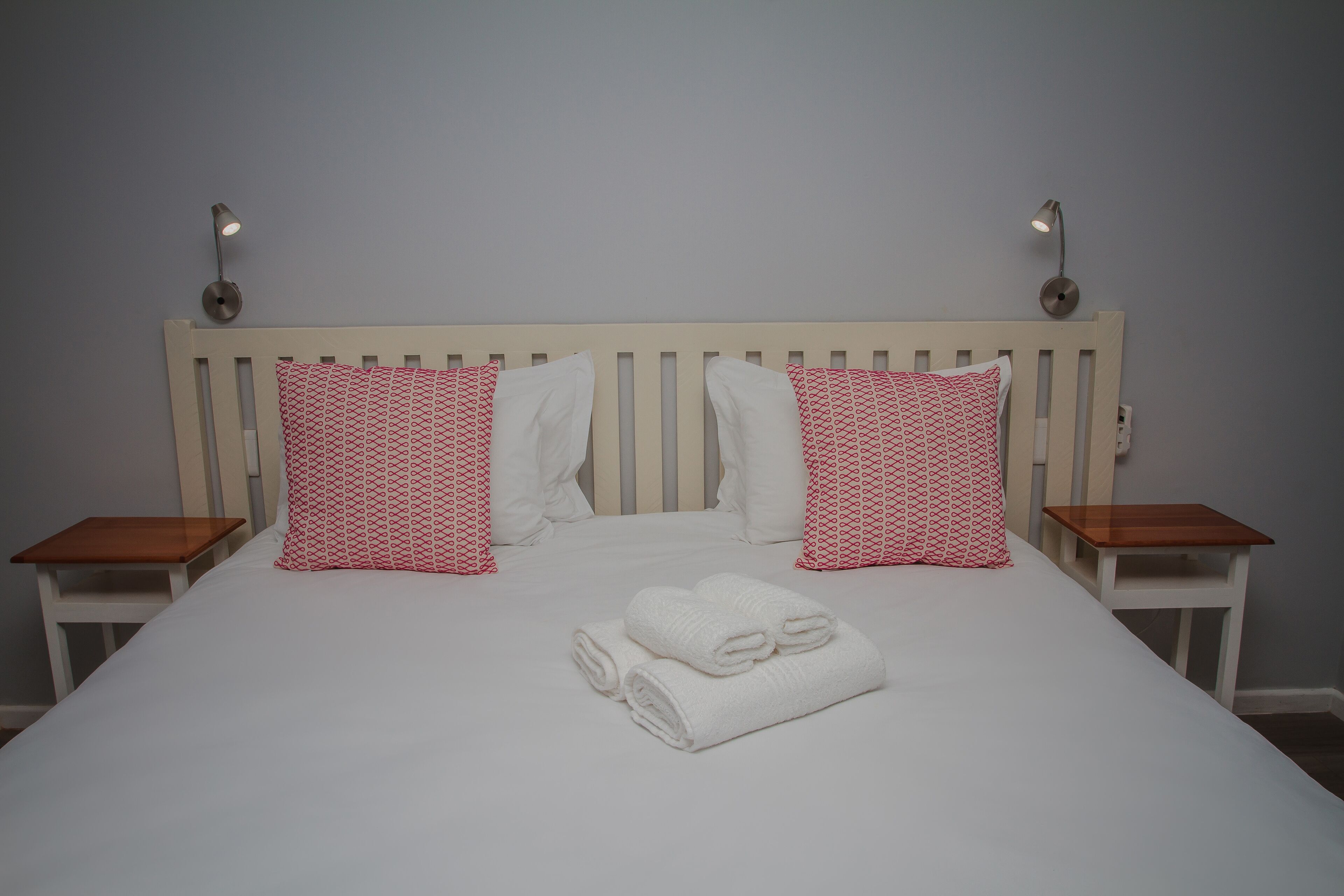 A neatly made bed with white bedding and red patterned pillows, two bedside tables, and wall-mounted lamps.