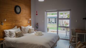 Habitación de hotel con cabecera de madera, cama con ropa de cama blanca, mesita de noche, escritorio con silla y vista al exterior a través de una puerta corrediza de vidrio.