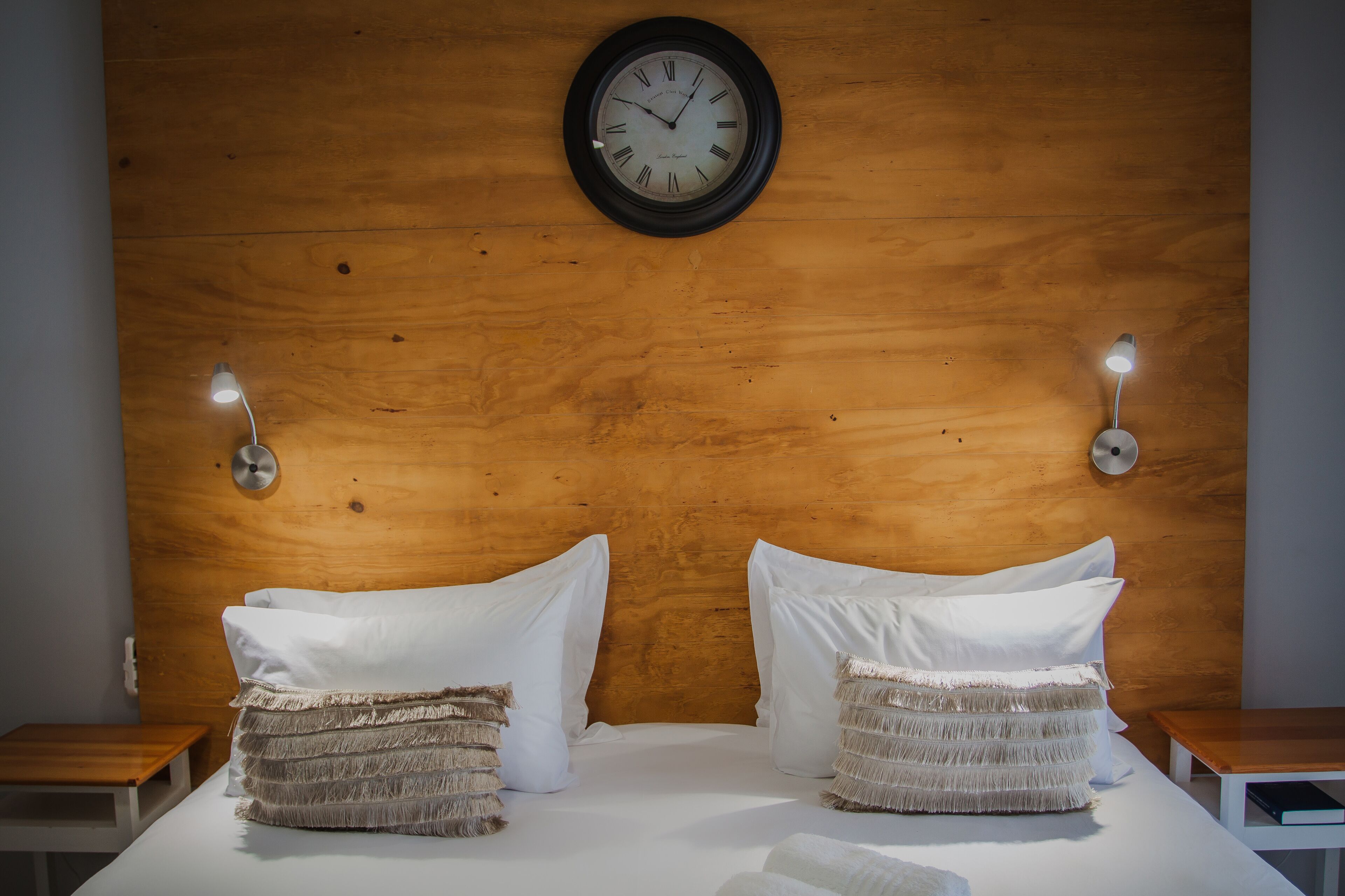 A neatly made bed with white linens and two textured pillows, a wooden headboard, a wall-mounted clock, and two bedside lamps.