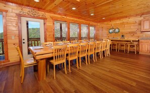 A wooden cabin with a dining area featuring a long table and chairs, a kitchen area, and large windows offering a view of the outdoors.