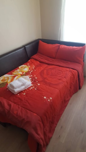 Una cama individual con una colcha roja y almohadas, una toalla blanca doblada y una funda de almohada estampada con motivos florales.