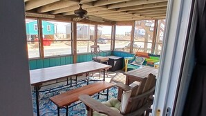 Ein überdachter Außenbereich mit Holztischen und -stühlen, einem Deckenventilator und Blick auf Strand und Gebäude im Hintergrund.
