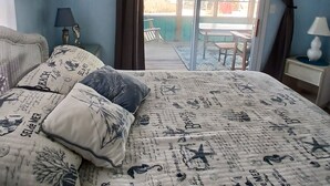 Ein Schlafzimmer mit Bett, Deckenventilator, Fenster mit Vorhängen, Nachttisch und Blick ins Freie.