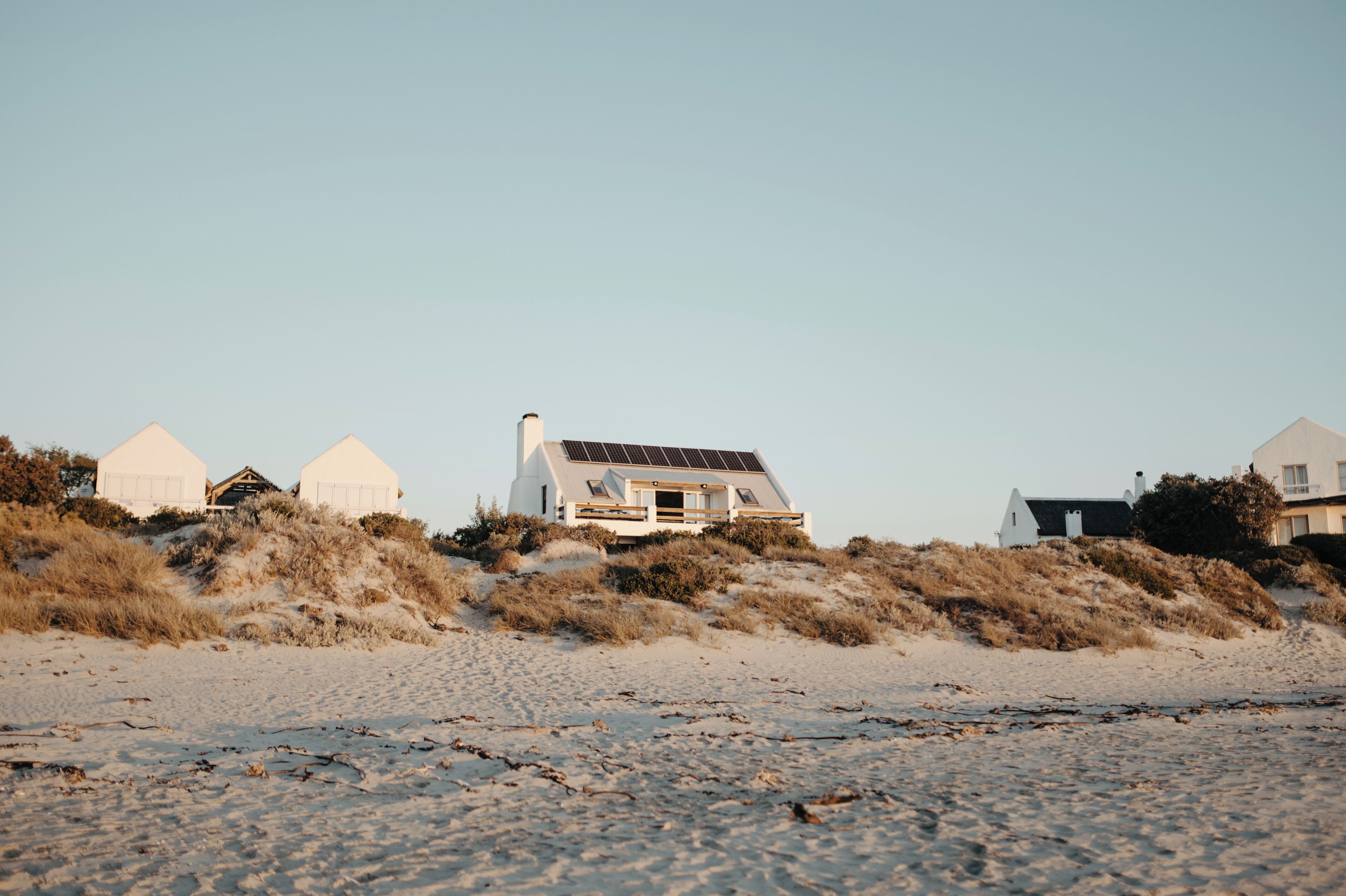 A coastal landscape with a row of white houses on a sandy beach, featuring a prominent house with a solar panel roof.