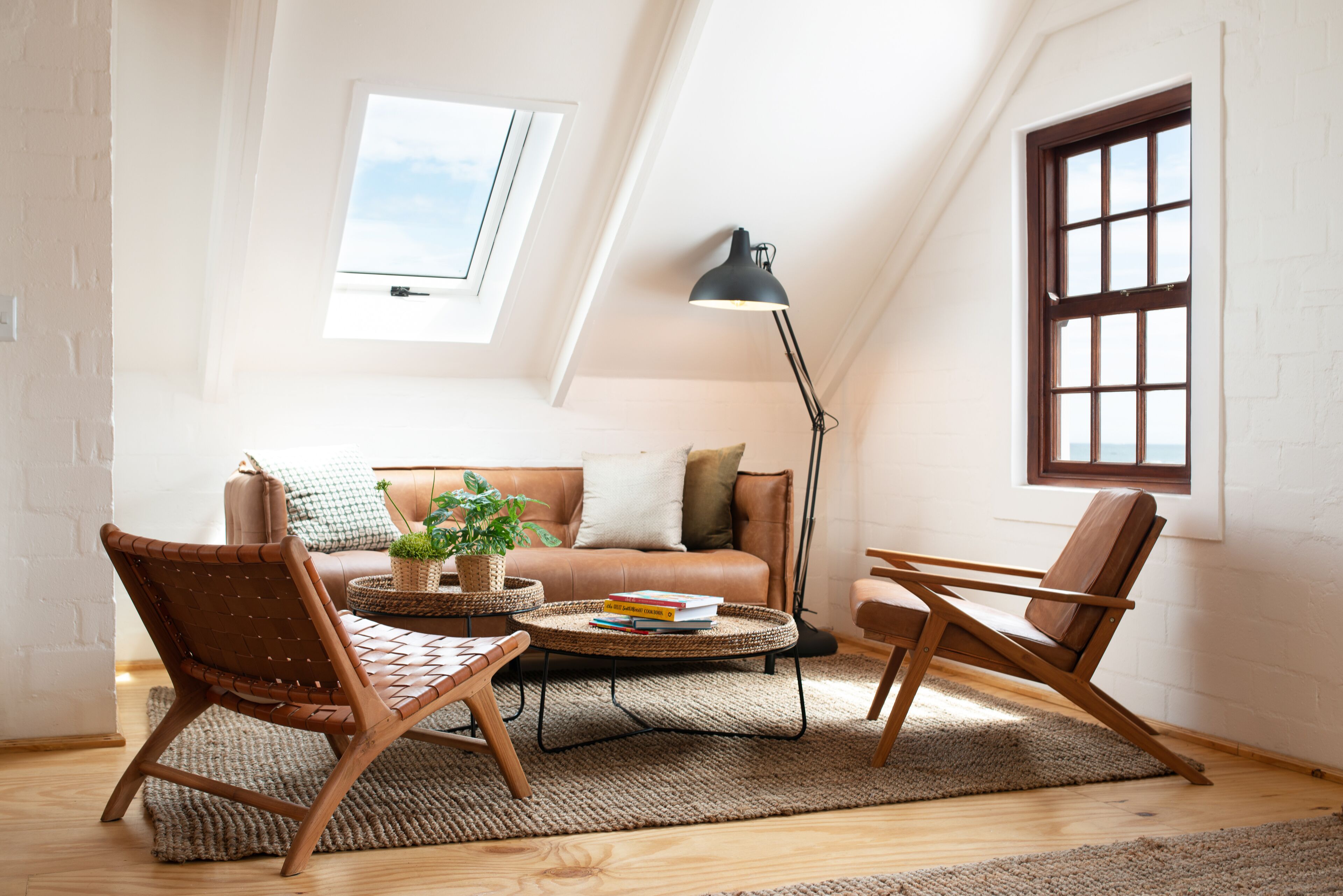 A cozy loft bedroom with a sloped ceiling, a large window, a floor lamp, a sofa, and two chairs.