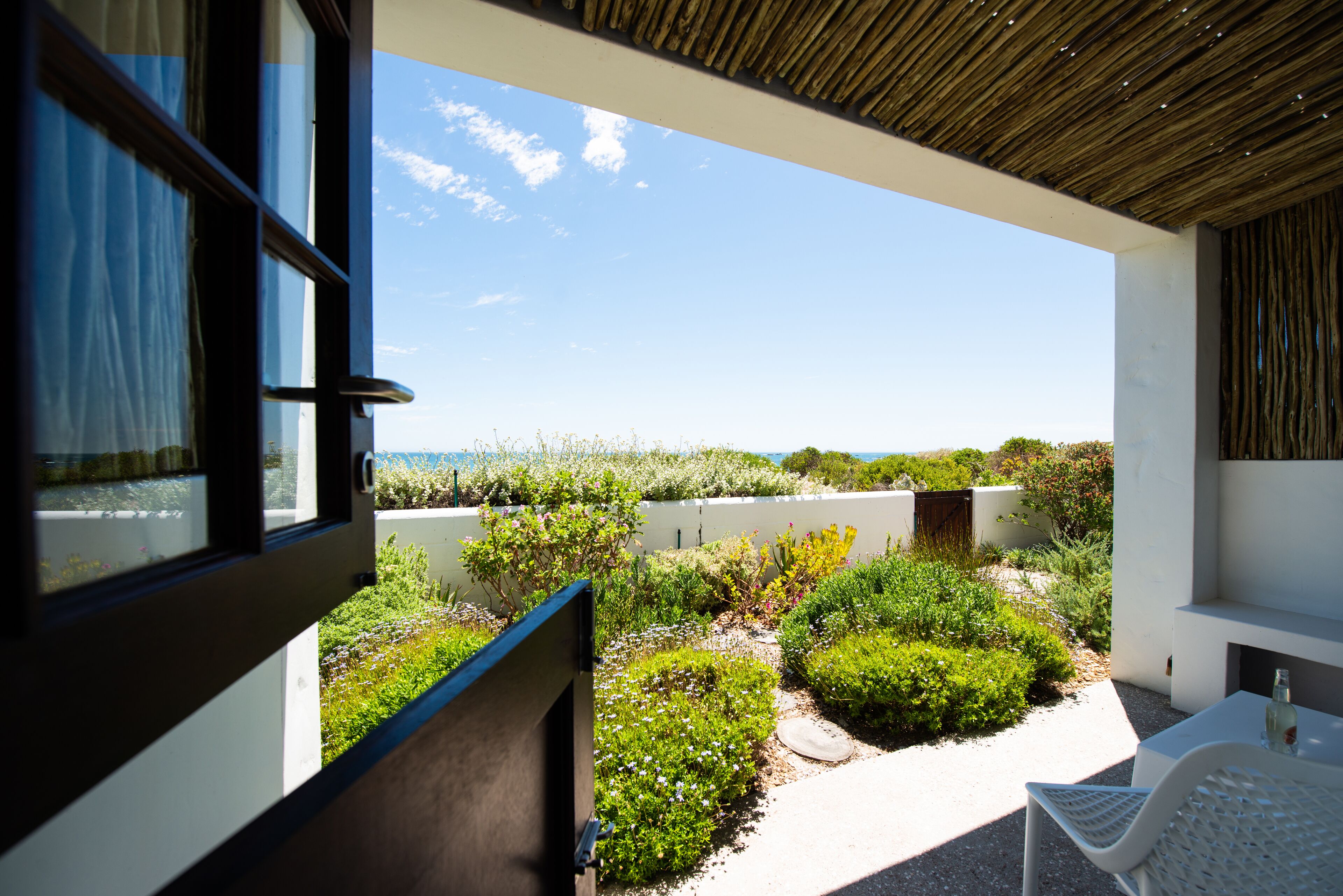 An open window with a view of a landscaped garden and a white table with a bottle.