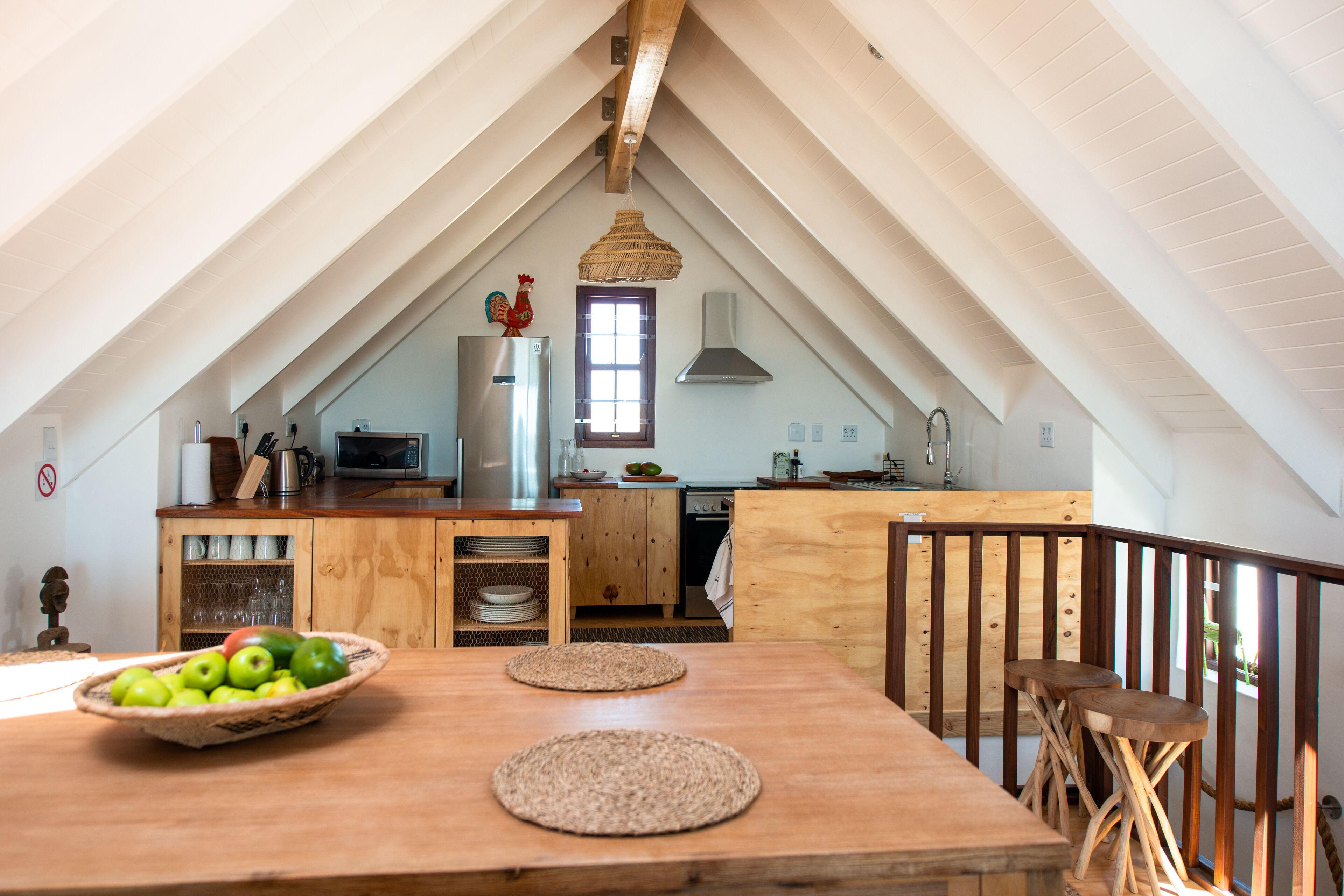 An attic kitchen with wooden beams, a dining table with a woven placemat, and a bowl of fruit.