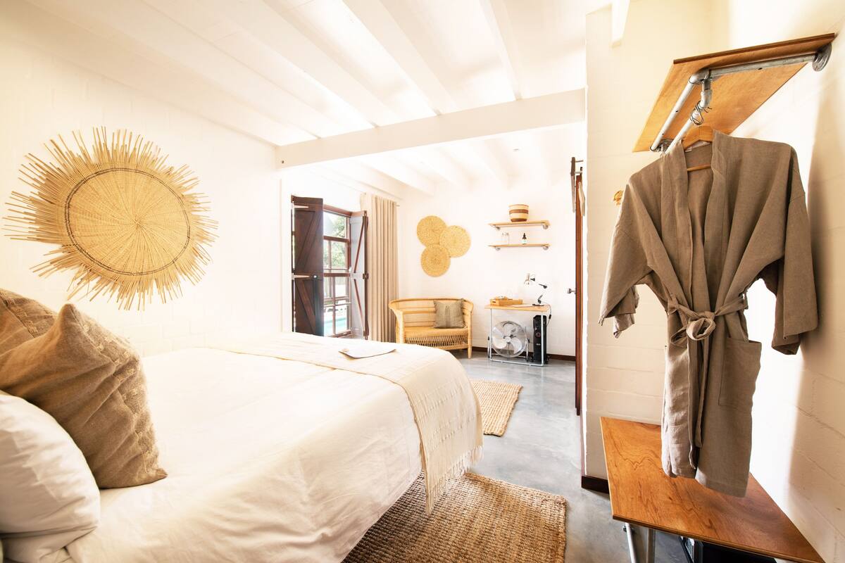 A bedroom with a large bed, a wooden table, a robe hanging on a rack, and a wall-mounted fan.