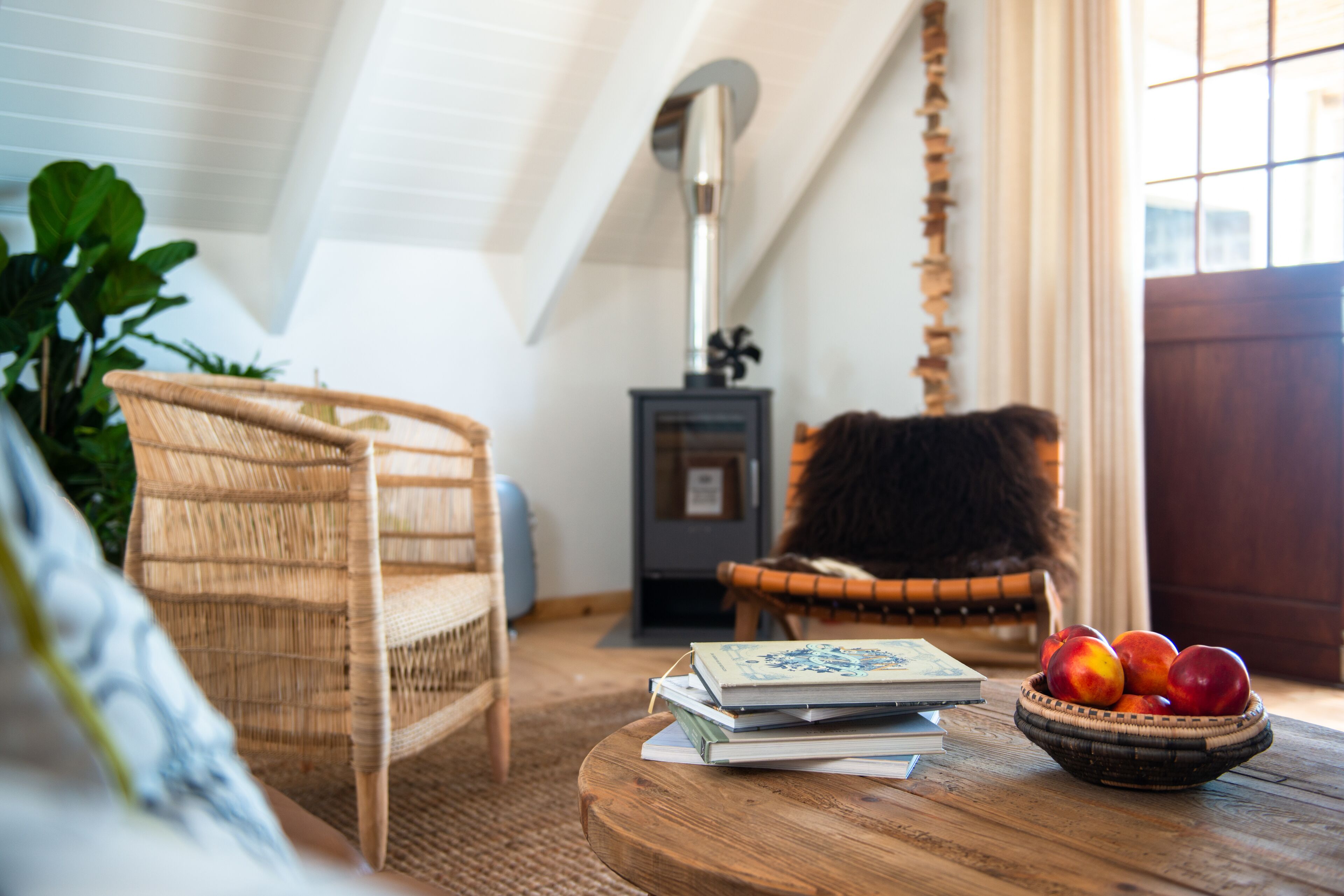 A cozy room with a wicker chair, a small table with books and a bowl of fruit, a fireplace, and a large window.