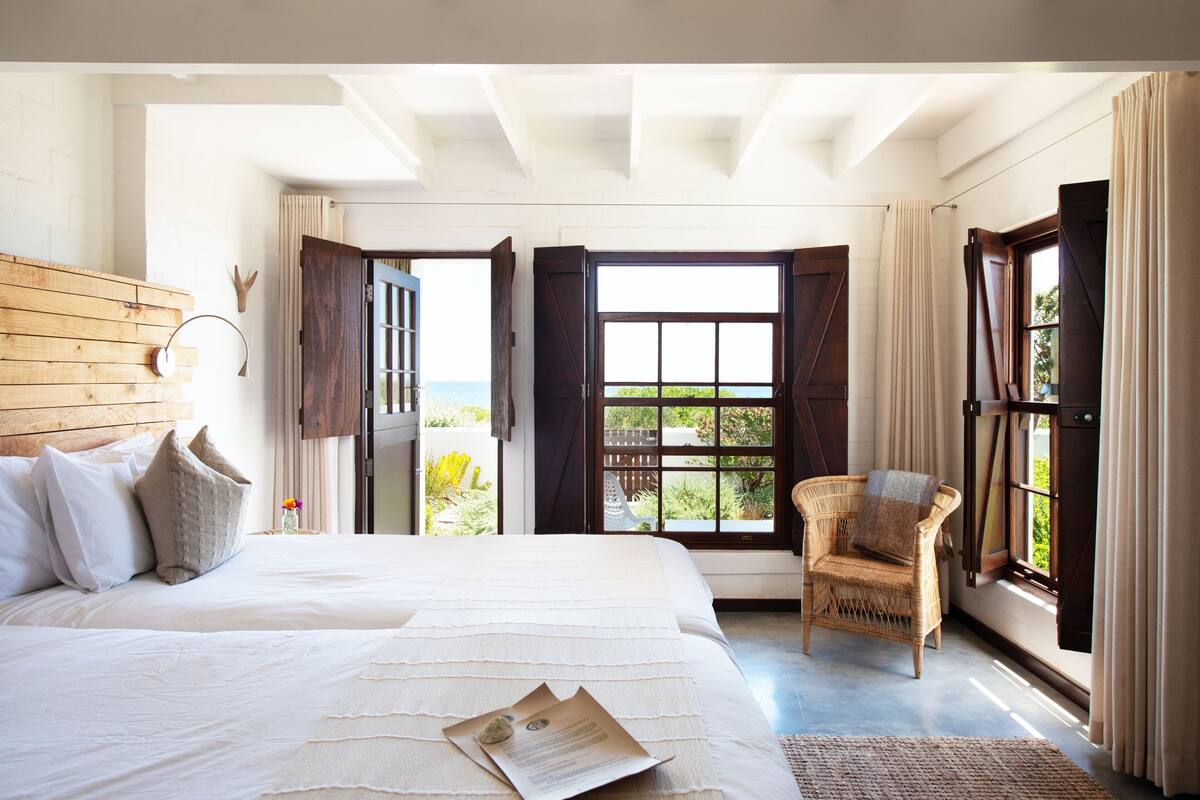 A bedroom with a large bed, a wicker chair, and a view of the outdoors through open doors.