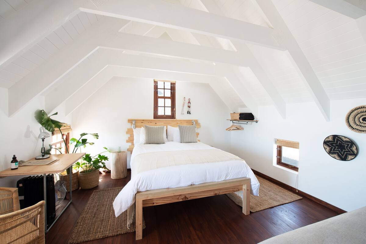 A bedroom with a sloped ceiling, a wooden bed, a desk with a lamp, and a potted plant.