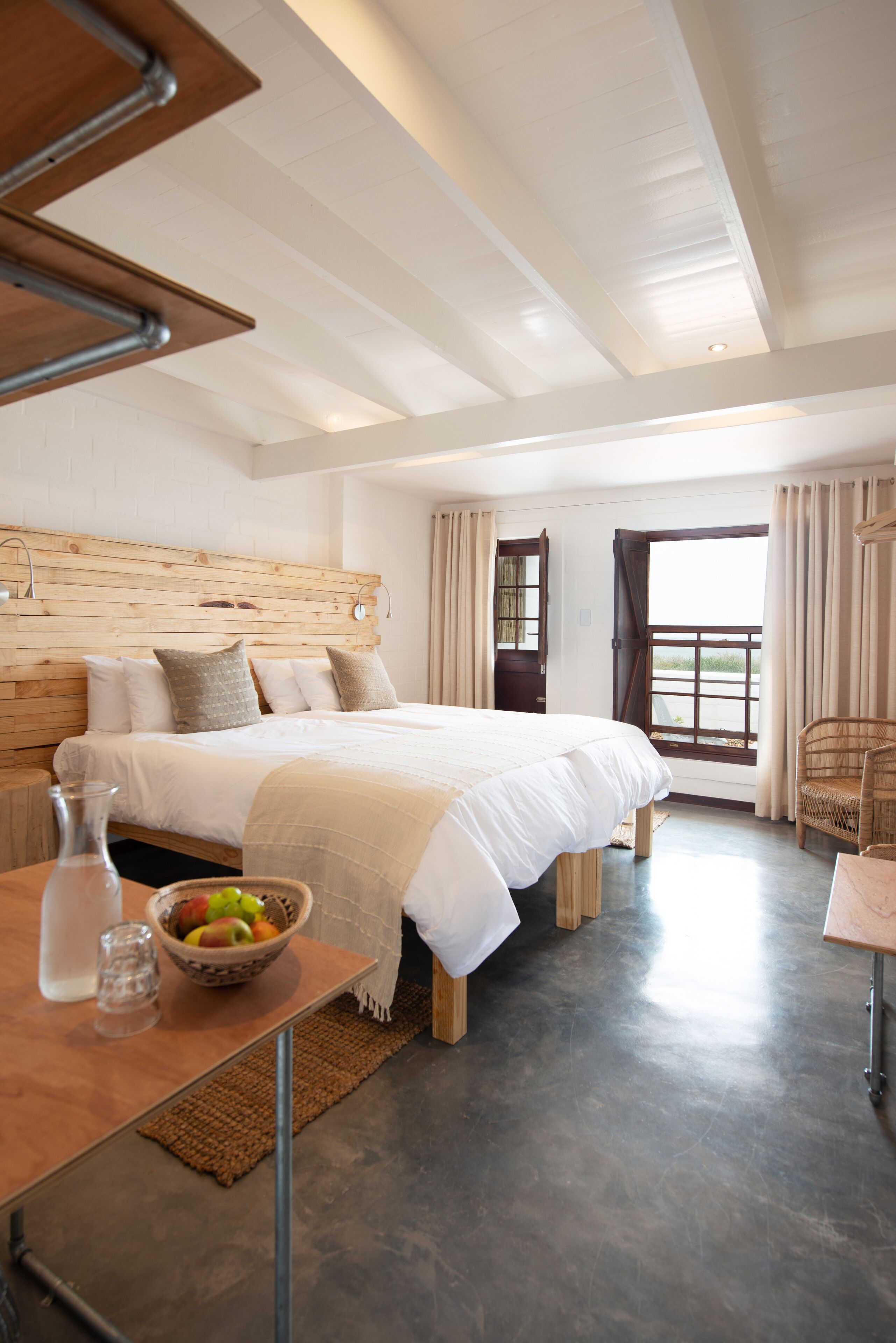 A modern bedroom with a wooden headboard, a large bed, a wooden table with a fruit bowl, and a view of the outdoors through a window.