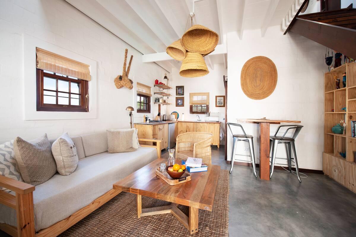 A modern living room with a wooden sofa, a coffee table with a bowl of fruit, and a kitchen area with wooden cabinets and a dining table.