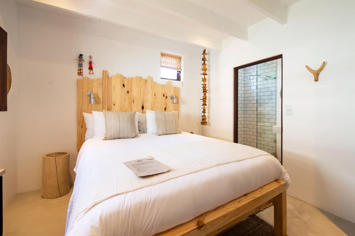 A bedroom with a wooden headboard, a bed with white bedding, a small wooden stool, and a window with blinds.
