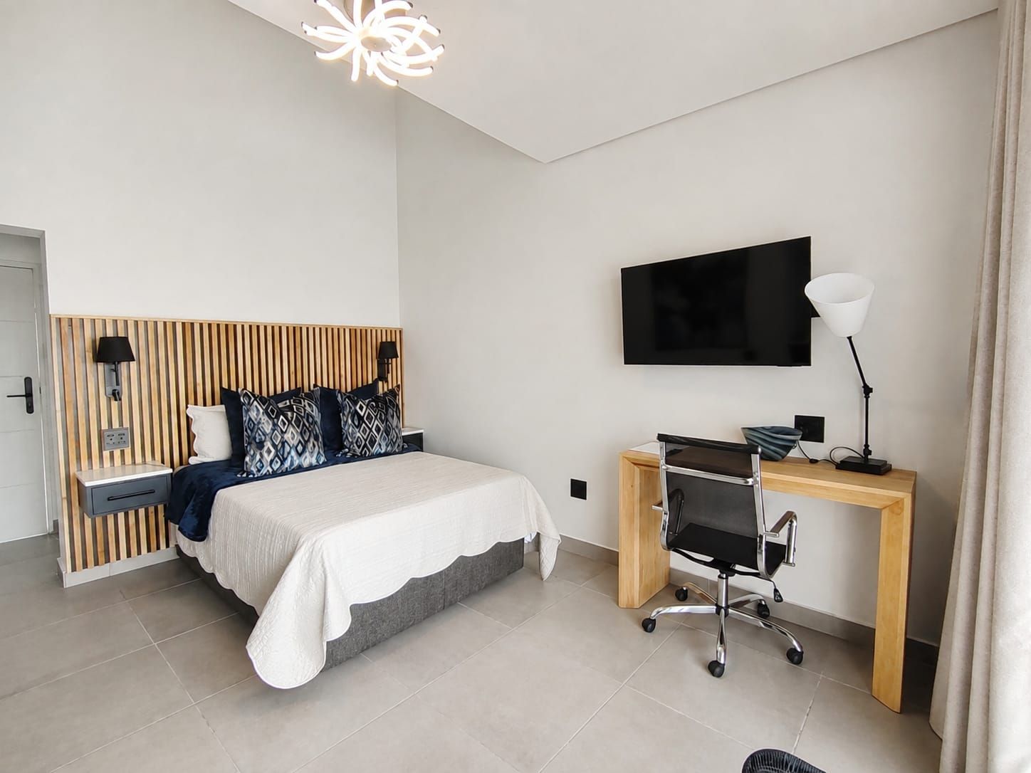 A modern bedroom with a bed, bedside table, lamp, desk, chair, and a mounted TV.