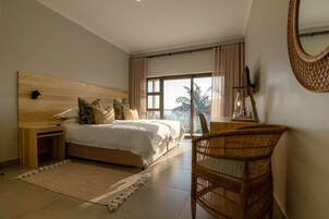 A bedroom with a wooden headboard, a large bed with white bedding, a wicker chair, a bedside table with a lamp, and a view of palm trees through a large window.