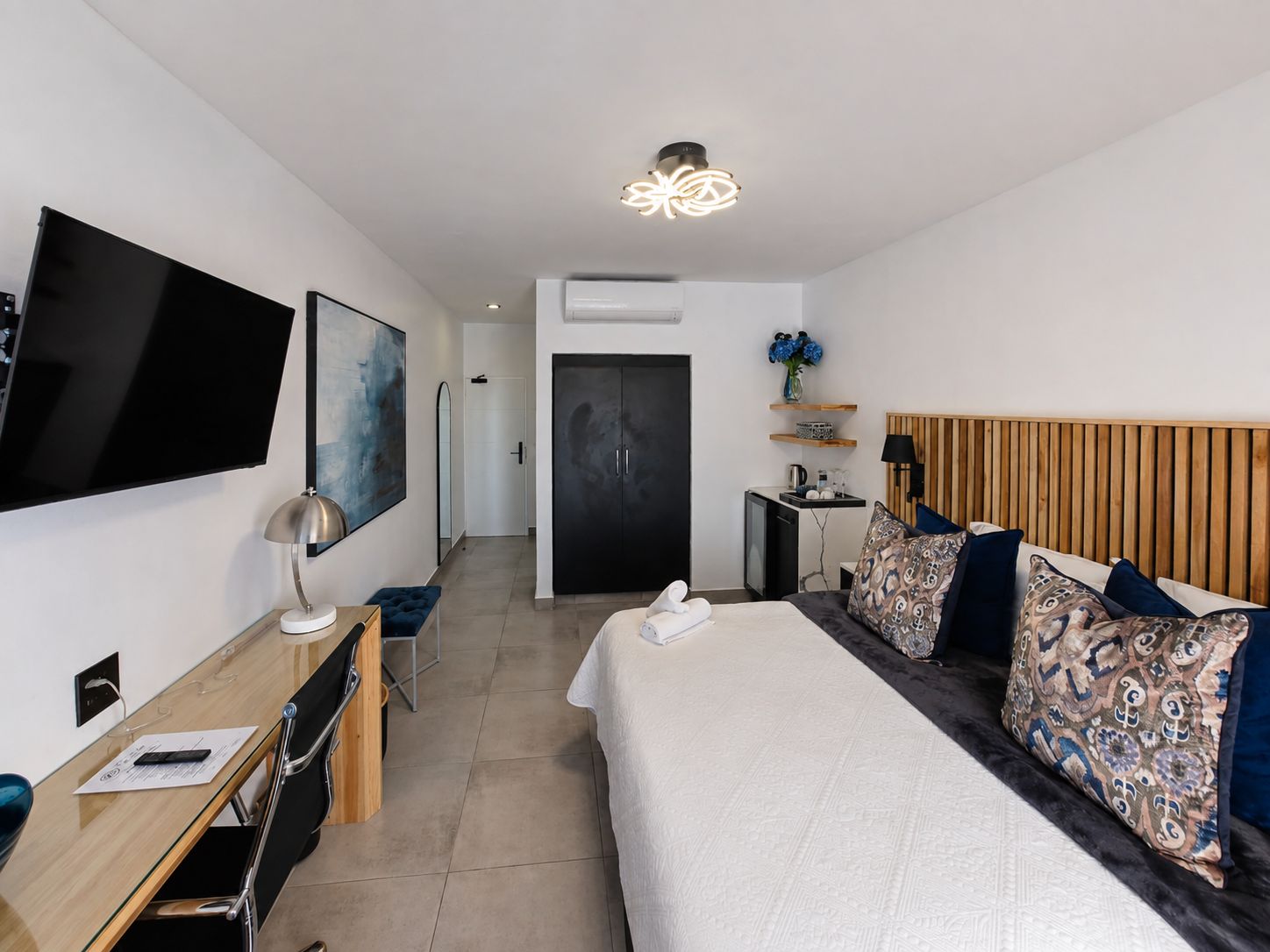 Habitación de hotel con cama, escritorio con silla, televisor de pantalla plana, un cuadro, lámpara del techo y una puerta.