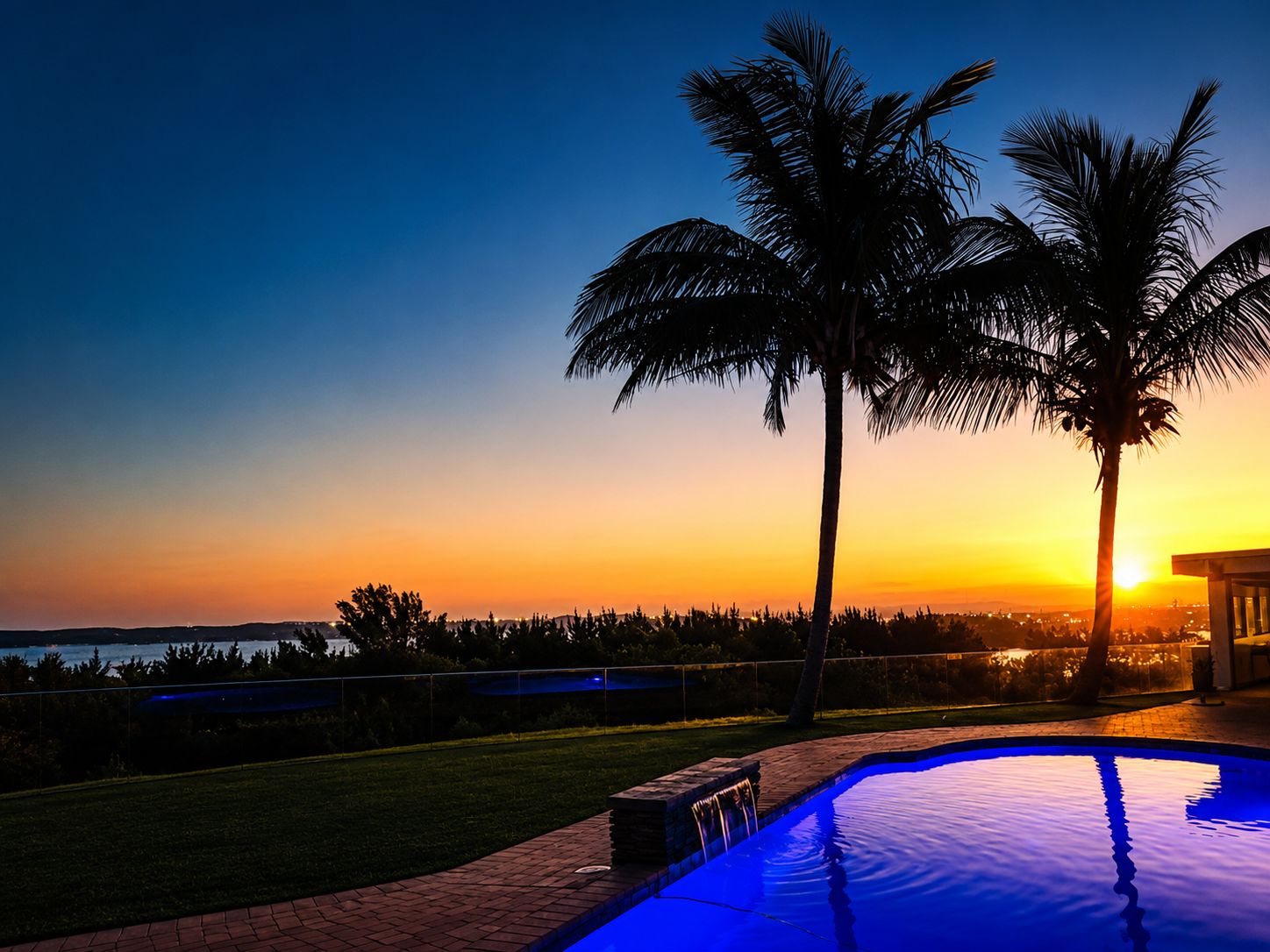 A swimming pool with a sunset view, palm trees, and a distant shoreline.