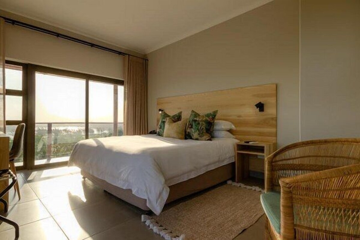Un dormitorio con una cama grande, cabecera de madera, mesita de noche, una silla de mimbre y vistas amplias a través de grandes ventanales.