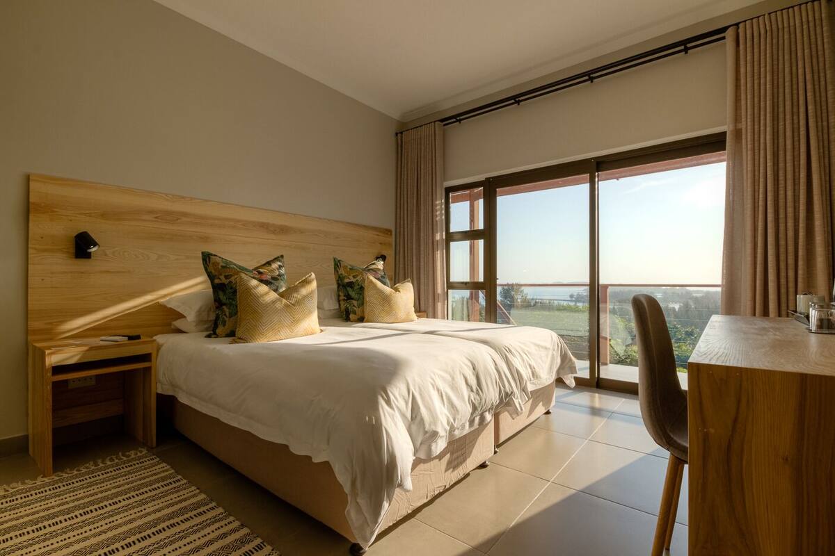 A modern bedroom with a large bed, a wooden headboard, a bedside table, a desk, and a view of the outdoors through a large window.