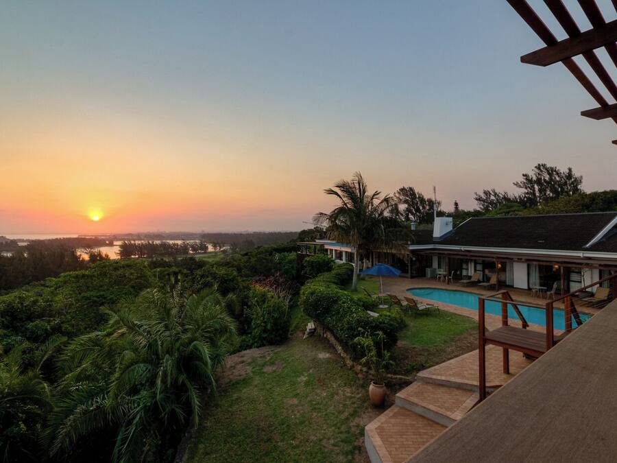 A house with a swimming pool, surrounded by lush greenery and a view of the sunset over a body of water.